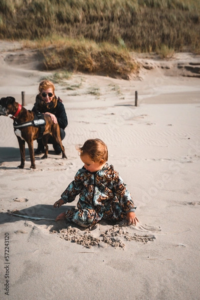 Fototapeta Kleinkind spielt im Sand am Starn, im Hintergrund hockt Mutter mit Boxer Hund am Rand der Dünen