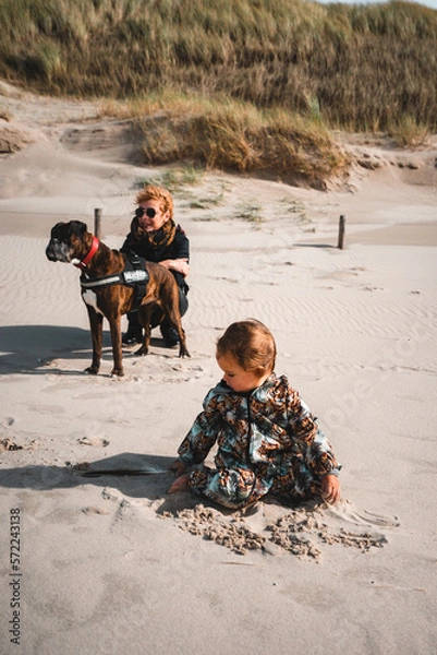 Fototapeta Kleinkind spielt im Sand am Starn, im Hintergrund hockt Mutter mit Boxer Hund am Rand der Dünen