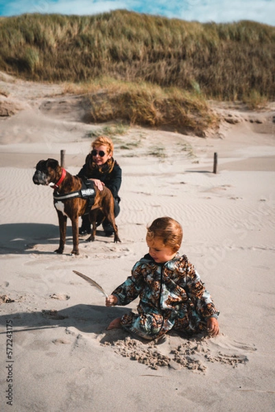 Fototapeta Kleinkind spielt im Sand am Starn, im Hintergrund hockt Mutter mit Boxer Hund am Rand der Dünen