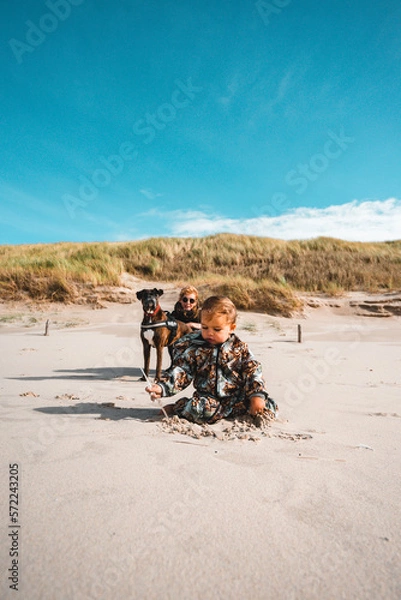 Fototapeta Kleinkind sitz am Strand im Vordergrund, dahinter hocken Mutter und Hund und schauen zu der Tochter die im Sand spielt