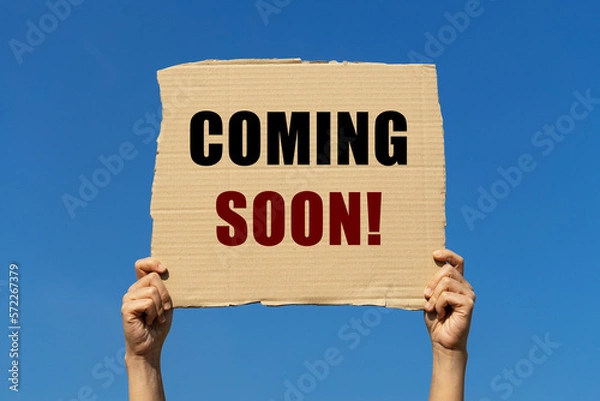 Fototapeta Coming Soon text on box paper held by 2 hands with isolated blue sky background. This message board can be used for business concept about something interesting will be available soon.