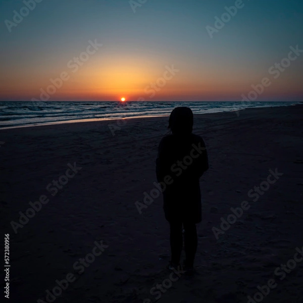 Fototapeta A young woman standing on a sea shore looking into a beautiful and colorful sunset on a calm as ice sea