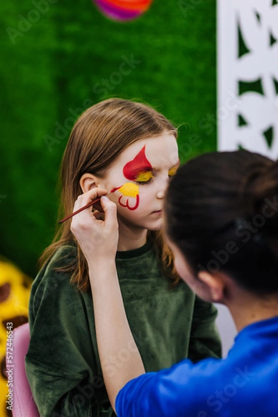 Fototapeta A professional make-up artist, artist paints with a brush on her face with face paints, drawing, children's makeup for a little girl model, a child. Photography, art, concept, lifestyle.