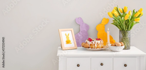 Fototapeta Easter cake, eggs and decorations on table near light wall in room