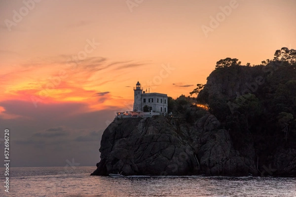 Obraz A splendid sunset by the historic lighthouse, Faro di Portofino, at Portofino, Liguria, Italy. A photo taken from the sea below with the rocky mountains and trees in the background.