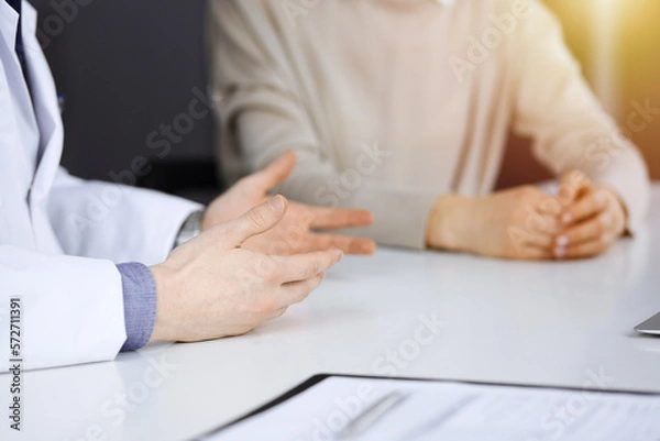 Fototapeta Unknown male doctor and patient woman discussing something while sittingin a darkened clinic, glare of light on the background. Close-up of hands