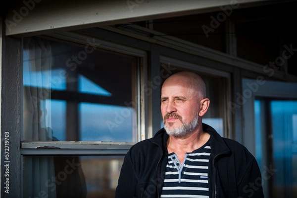 Obraz ein Mann um die 50 mit Bart steht unterm Vordach eines Holzhauses in schöner Landschaft und blickt in die Ferne.