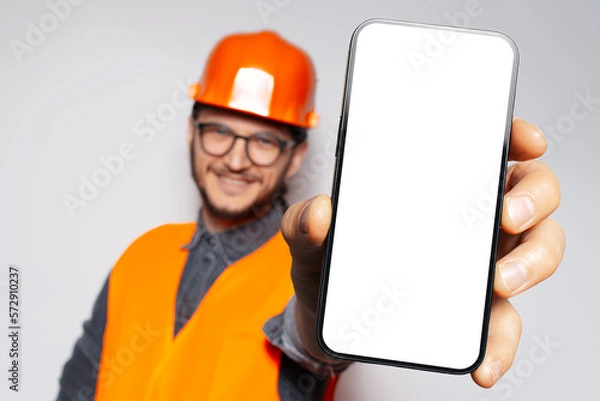 Fototapeta Big smartphone with blank on screen in hand on construction worker, smiling man. Close-up view. Mock up concept. White background.