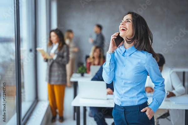 Fototapeta Smiling businesswoman using phone in office. Small business entrepreneur looking at her mobile phone and smiling. Beautiful smiling businesswoman using smarphone indoors, copy space.