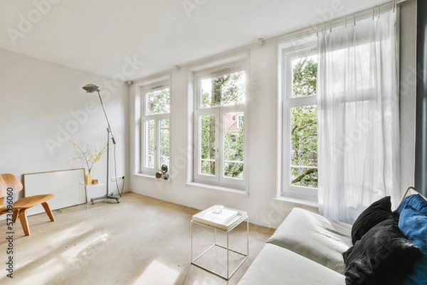 Fototapeta a living room with white walls and wood flooring, two large windows in the wall is covered with sheer curtains