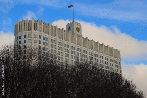 Fototapeta The White House of the Government of the Russian Federation with the Russian flag on the roof - against a blue sky with white clouds and tree branches in the foreground.