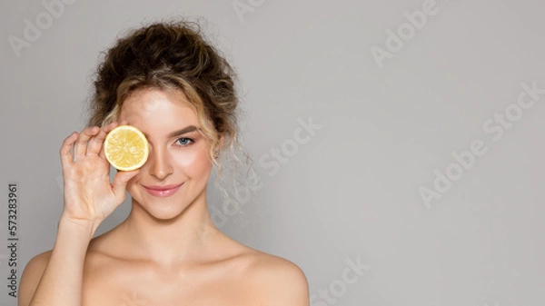 Fototapeta Beautiful woman holding lemon half and looking at camera, lady using vitamin c fruit acids for beauty care, copy space