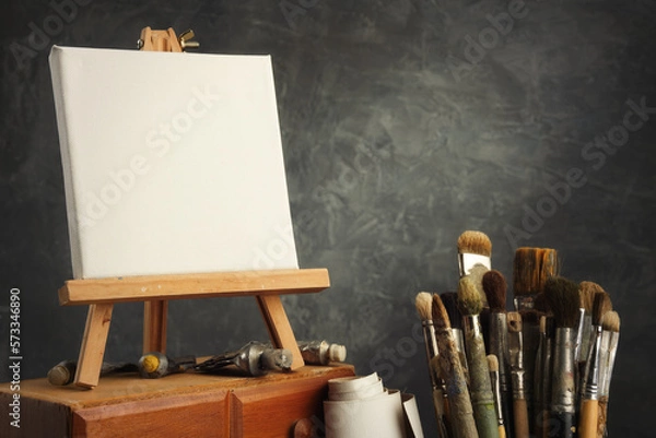 Fototapeta Artistic equipment in a artist studio: empty artist canvas on a wooden easel, paint tubes and paint brushes - used artistic paintbrushes for painting with oil or acrylic paints.