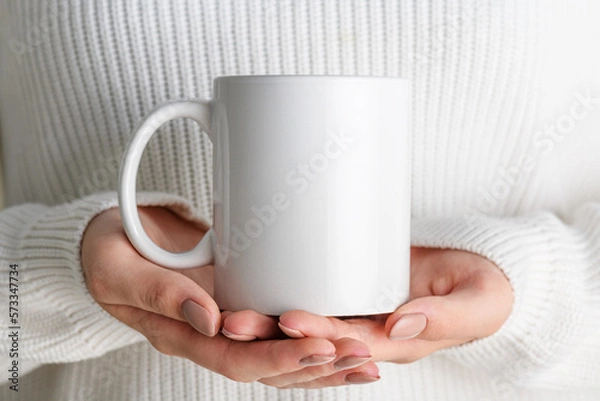 Obraz Female hands holding white mug mockup with blank copy space for your advertising text message or promotional content. Girl in white sweater holding white porcelain coffee mug mock up, close up