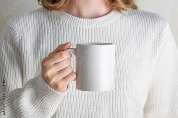 Obraz Female hands holding white mug mockup with blank copy space for your advertising text message or promotional content. Girl in white sweater holding white porcelain coffee mug mock up, close up