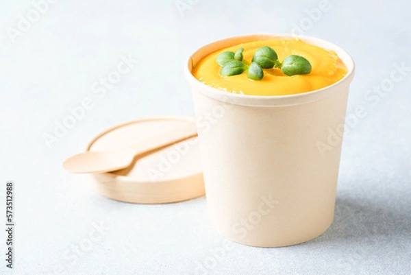 Fototapeta Creamy pumpkin fall soup in craft paper food container against light gray background. Butternut squash cream soup garnished with seasonings. Food delivery and soup to go. Mockup