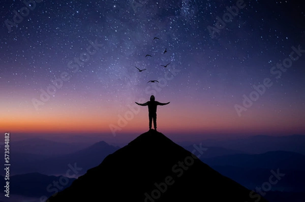 Fototapeta Silhouette of young man standing alone on top of mountain and raise both arms praying and free bird enjoying nature on beautiful night sky, star, milky way background. Demonstrates hope and freedom.