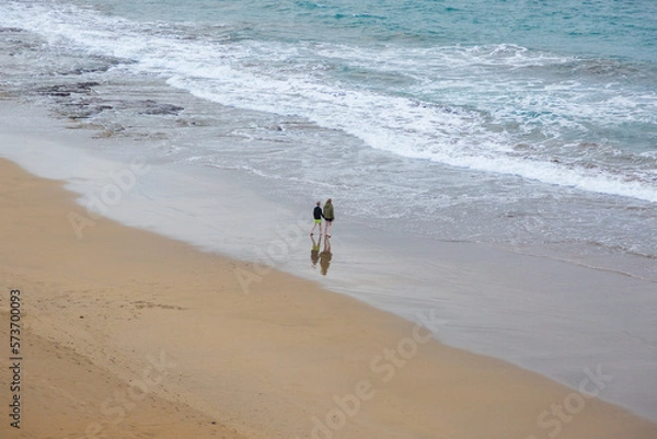 Fototapeta Woman and a child holding hands, standing on the beach, looking into the ocean on overcast cold day wearing jackets and shorts.