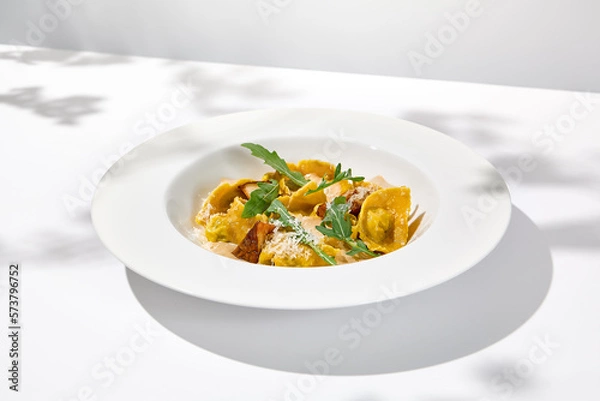 Fototapeta Italian ravioli with meat and mushroom sauce on white plate. Meat tortellini with creamy sauce in summer menu with shadows of tree leaves Ravioli stuffed beef in elegant style