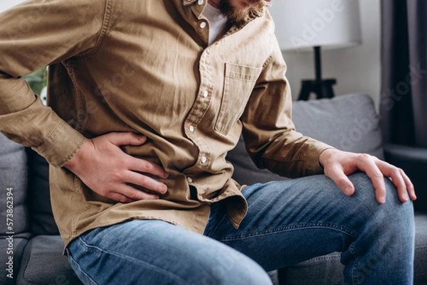 Obraz Sad young man sitting on couch at home, suddenly feeling strong stomach ache, gastritis problem. Upset male 30s squeezing belly with hand because of abdominal pain. Guy suffering from stomach ache
