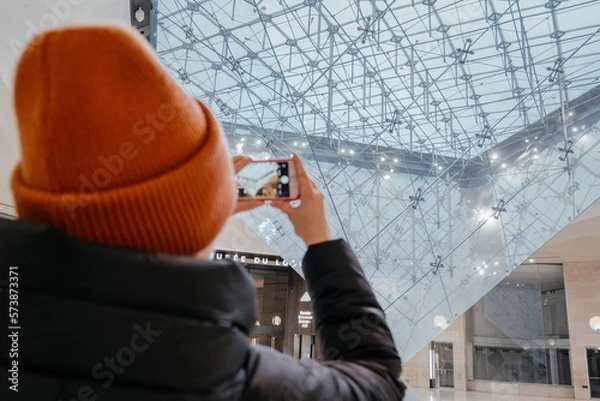 Obraz Young woman taking a photo. Inverted pyramid in the shopping mall 'Carrousel du Louvre' with people in Paris, France. Louvre museum hosts one of the biggest art collection in the world.