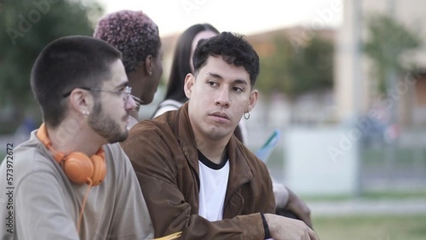 Obraz Concentrated diverse young male friends in casual clothes with headphones sitting on bench while having conversation on street