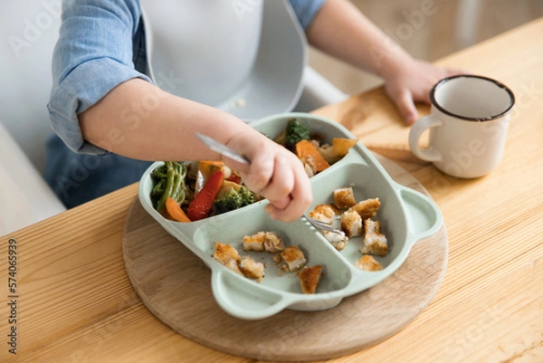 Obraz Top view of how a toddler boy pricking food with a fork from a special children's plate. Concept of self-feeding. BLW. Child without the help of adults eats healthy vegetables with meat. close-up
