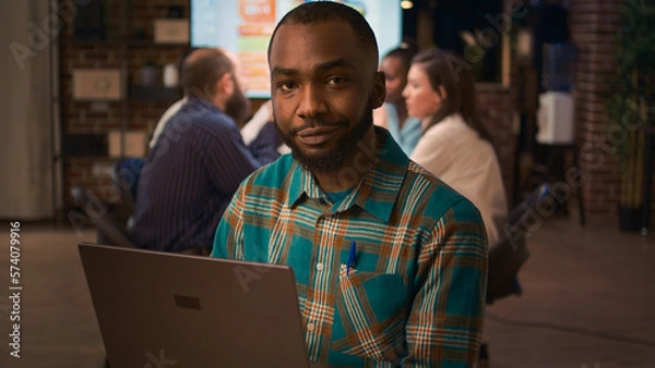 Fototapeta African american smiling employee working on laptop portrait, slow motion, front view. Office worker holding portable computer, looking at camera medium shot, business meeting in background