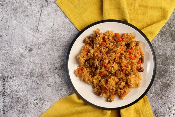 Fototapeta Homemade pilaf with rice, meat, carrots and onions on a white round plate on a dark gray background. Top view, flat lay