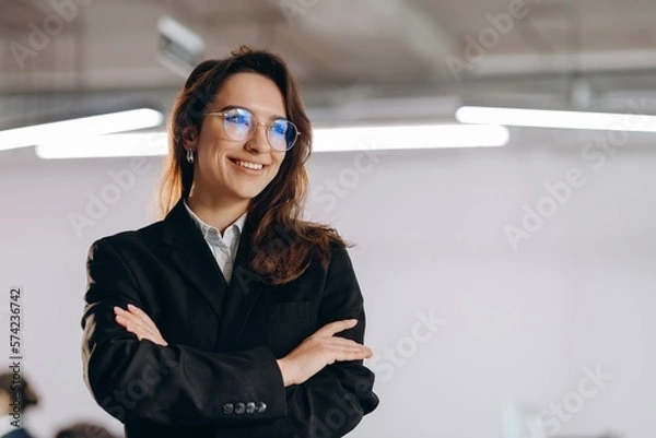 Fototapeta Portrait of smiling manager girl wearing glasses and black suit. Happy businesswoman standing with arms folded and looking away