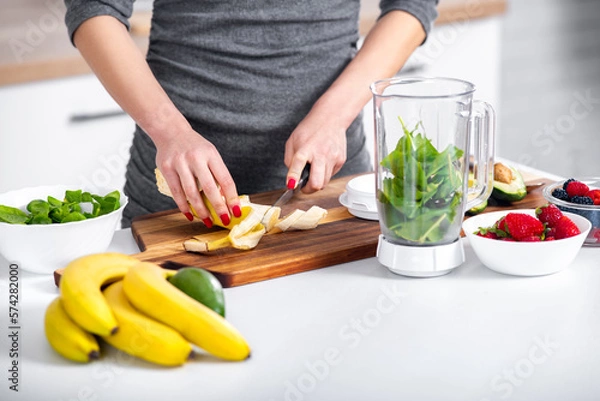 Fototapeta Woman with red nails chopping banana when preparing healthy smoothie. Raw drink made of banana, berries, avocado and spinach.