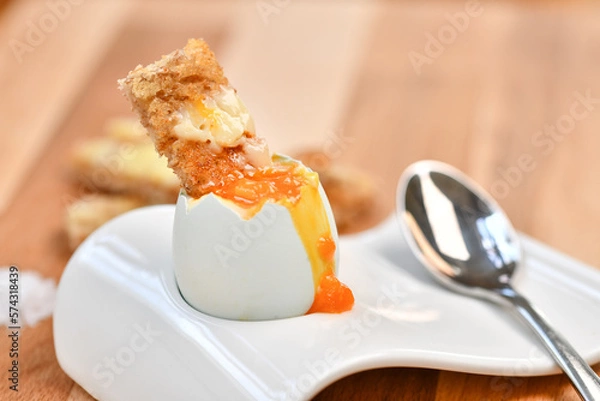 Fototapeta a single boiled egg in an egg cup, broken open showing yolk and with a slice of toast inserted with a spoon on a wooden surface