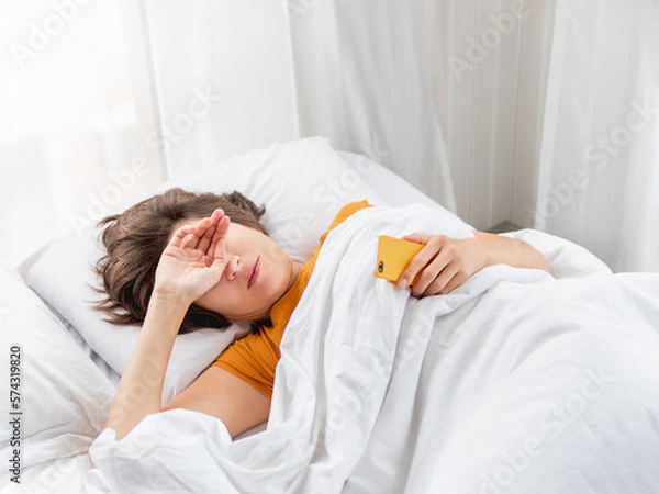 Fototapeta Sleepy woman is lying in bed, completely covered with white blanket. Smartphone used as alarm clock. It's hard to wake up early in morning. Woman does not get enough sleep.