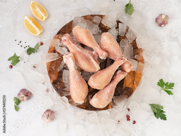 Fototapeta frozen raw chicken legs with vegetables on the ice on wooden board on the.gray rock background