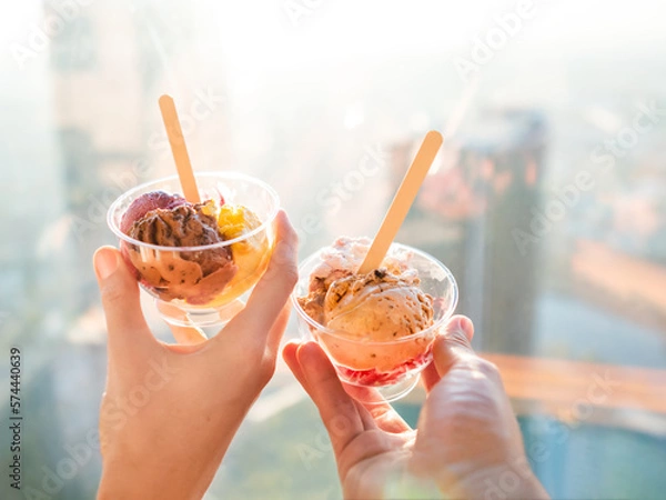Fototapeta Man and woman holds plastic bowls with colorful balls of ice-cream. Cold dessert for sunny days. Different gelato with wooden spoons. Romantic date in cafe.