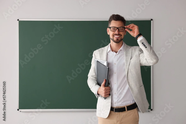 Fototapeta Happy teacher with glasses and stationery at blackboard in classroom. Space for text