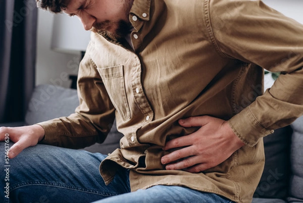 Fototapeta Upset male squeezing belly with hand because of abdominal pain. Unhappy young man sit on grey couch at home, suddenly feeling strong stomach ache, gastritis problem. Guy suffering from stomach ache