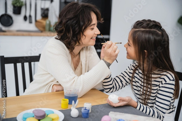 Fototapeta Side view of cheerful mother and child holding paintbrushes near Easter eggs and macaroons at home.