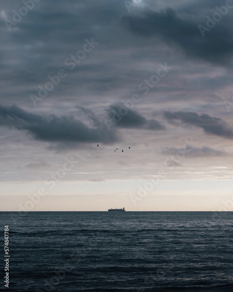 Obraz In the vast expanse of the ocean, a lone cargo ship cuts through the waves, its silhouette a stark contrast against the moody twilight sky.