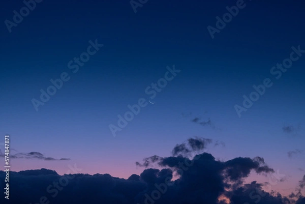Obraz The tranquil beauty of a small crescent moon suspended in a sea of blue and purple hues, peeking out from above the rolling clouds.