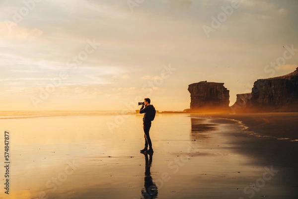 Obraz A lone photographer silhouetted against a blazing golden sunset, intently capturing the breathtaking scenery through his lens, at a stunning and secluded beach location.