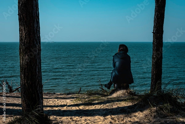 Fototapeta A lonely woman sitting between trees and watching to a calm and silent see meditating