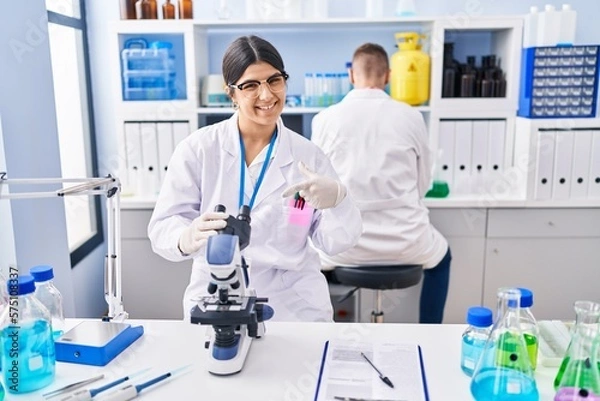 Fototapeta Young woman working at scientist laboratory smiling happy pointing with hand and finger