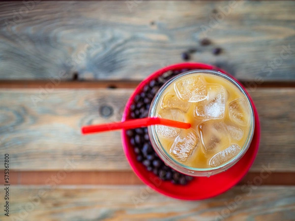 Fototapeta Ice coffee on a wood table with ice and red straw, coffe beans and red saucer on background. Cold summer drink with red tube