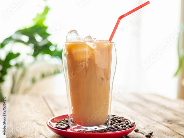Fototapeta a drink sitting on top of a red plate with coffee beans on top of a wooden table, plant on background. Iced latte in summer cafe