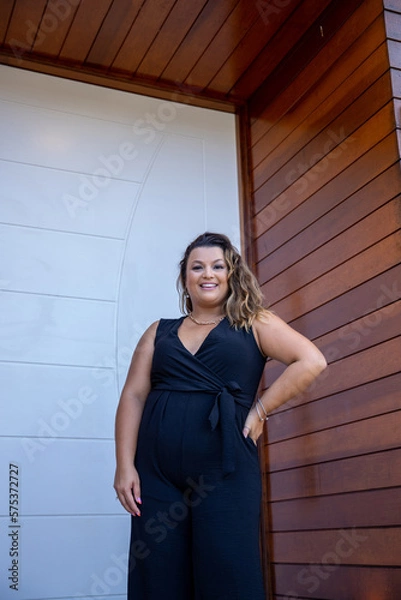 Obraz fat woman empowered in front of a house, successful businesswoman with hand on waist wearing elegant black jumpsuit, person in front of a house