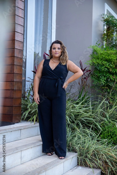 Obraz fat woman empowered in front of a house, successful businesswoman with hand on waist wearing elegant black jumpsuit, person in front of a house