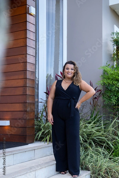 Obraz fat woman empowered in front of a house, successful businesswoman with hand on waist wearing elegant black jumpsuit, person in front of a house
