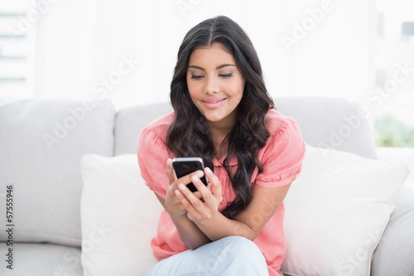 Obraz Content cute brunette sitting on couch holding smartphone