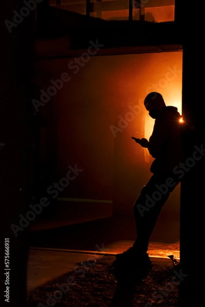 Fototapeta Silhouette of a masked man with a smartphone in orange smoke